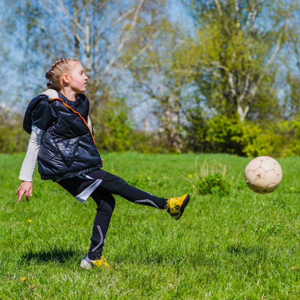 Mergaitė, vilkinti juodą sportinę aprangą ir geltonus sportinius batelius, spardo futbolo kamuolį žaliame lauke, apsuptame medžių.