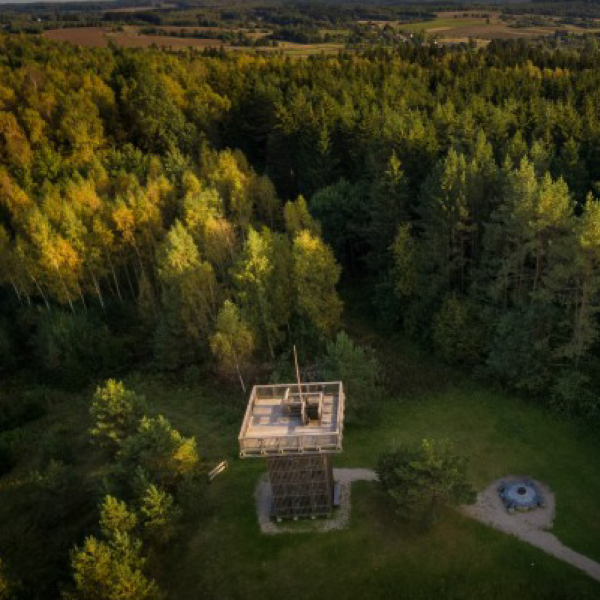 Apžvalgos bokštas miško viduryje, matomas iš oro, apsuptas medžių ir natūralios gamtos su tolumoje matomais laukais.